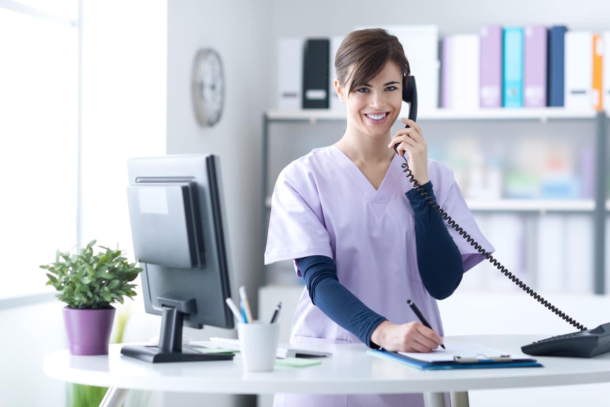A smiling dermatology front desk employee answering a phone call while taking notes